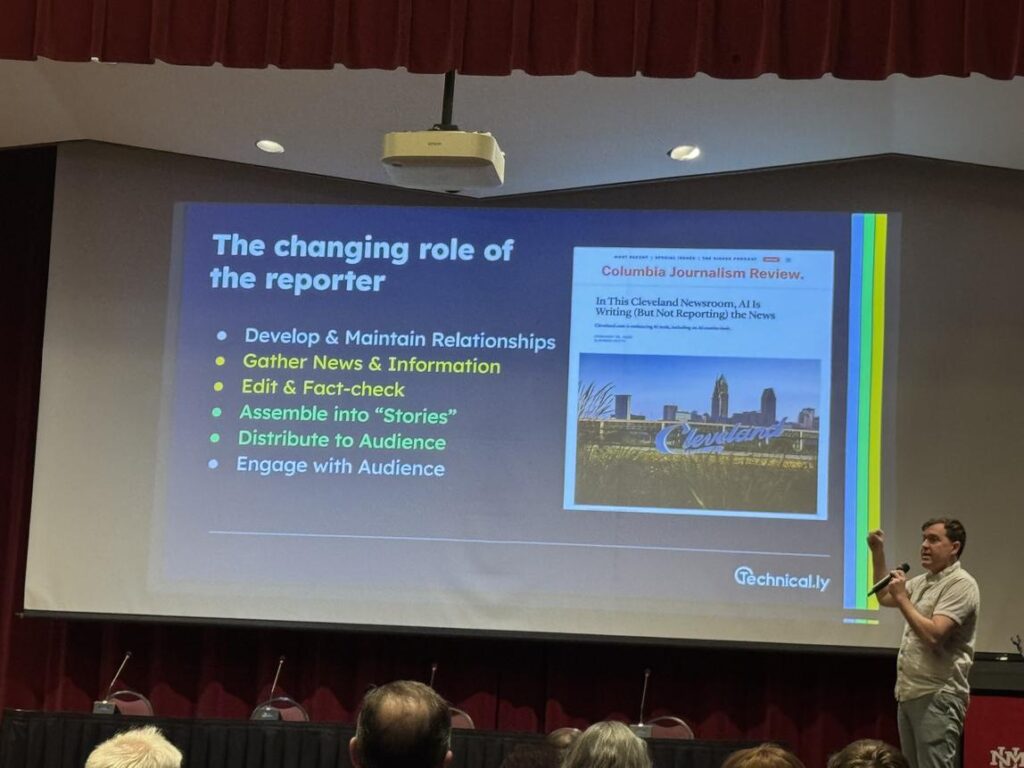 man speaking to crowd in front of projected screen